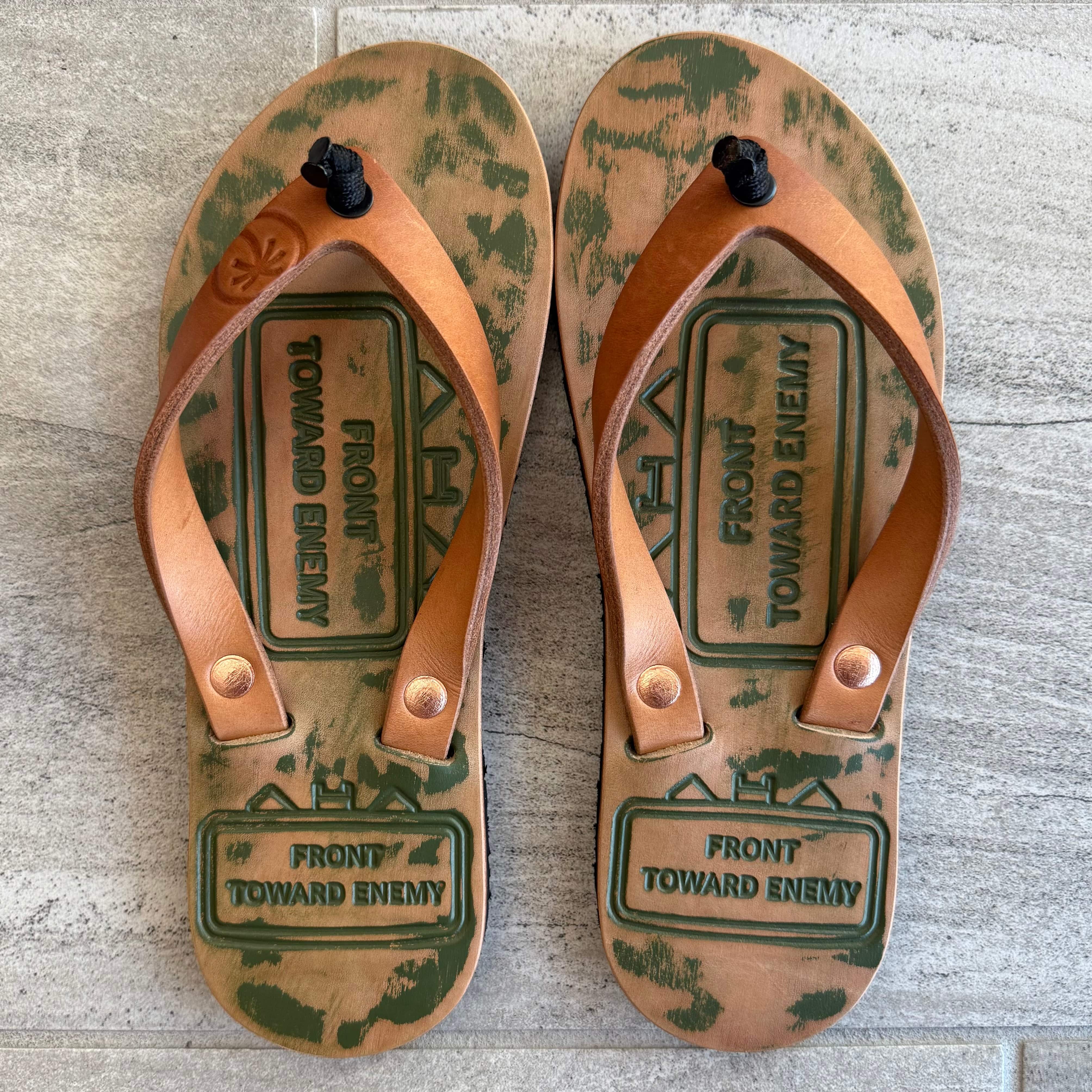 Toehold Claymore flip-flops with black straps and cushioned soles resting on a wooden surface.